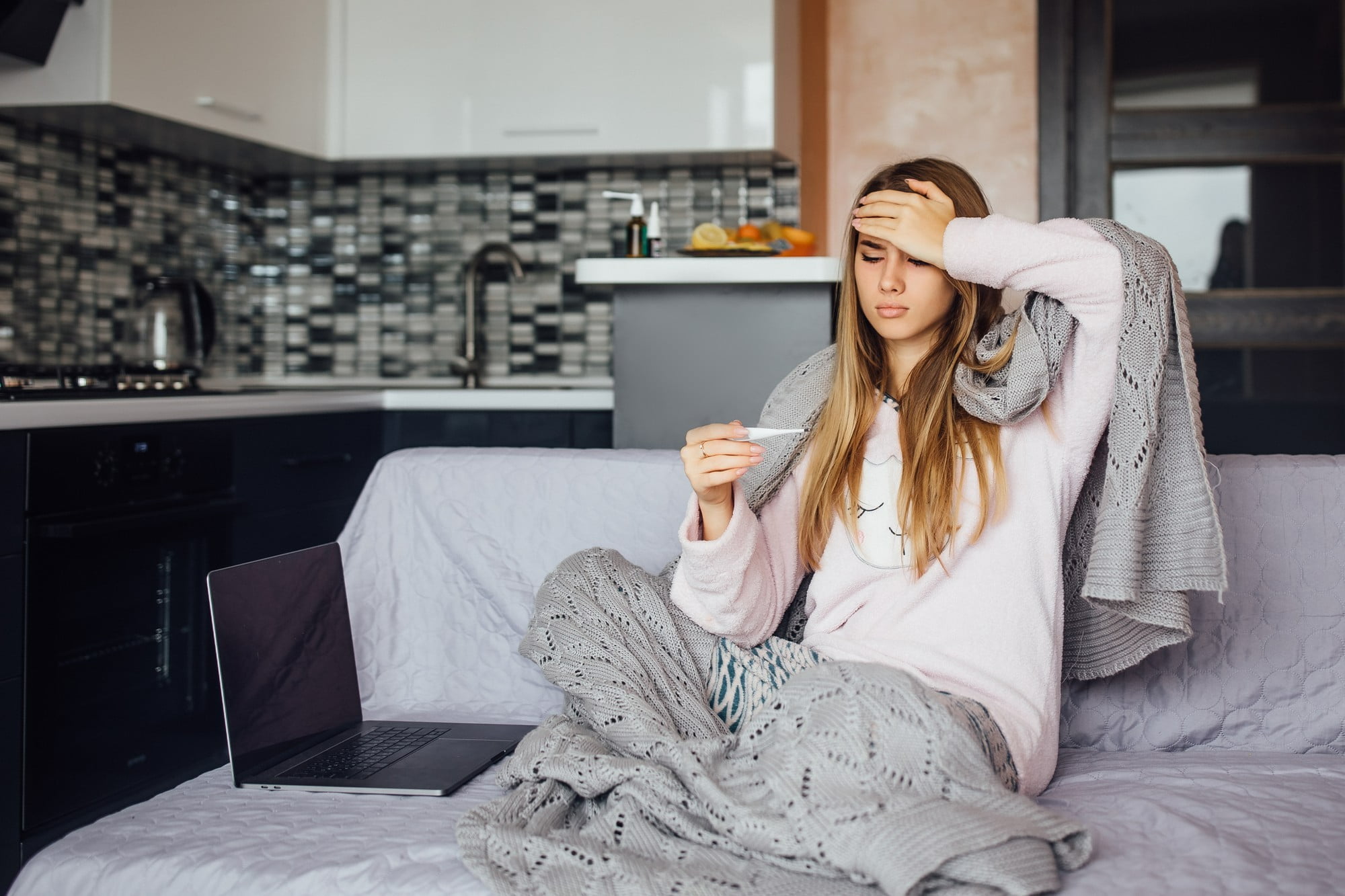 young woman sitting on couch with blanket taking temperature trying to stay healthy during the holiday season